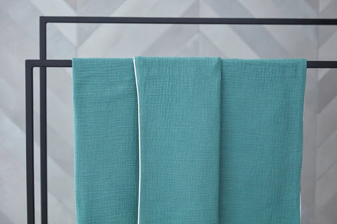 Ein blaugrünes Handtuch mit weißem Rand hängt fein säuberlich über einem schwarzen Metallgestell vor einem grauen, geometrisch gemusterten Hintergrund.