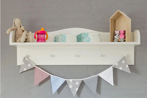 Auf einem weißen Regal stehen ein Plüschhase, ein rosa Trinkbecher, gestapelte Windeln und eine Spielzeugeisenbahn. Darunter hängen graue, rote, blaue und weiße Wimpelfahnen an einer grauen Wand.