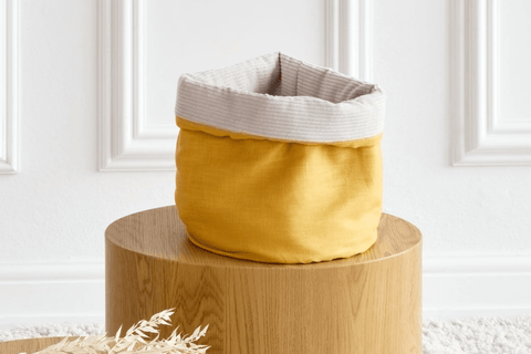 Ein gelb-beiger Aufbewahrungskorb aus Stoff mit gefaltetem Rand steht auf einem runden Holztisch vor einer weißen Wand mit Zierleisten. Im Vordergrund ist getrocknetes Pflanzendekor zu sehen.