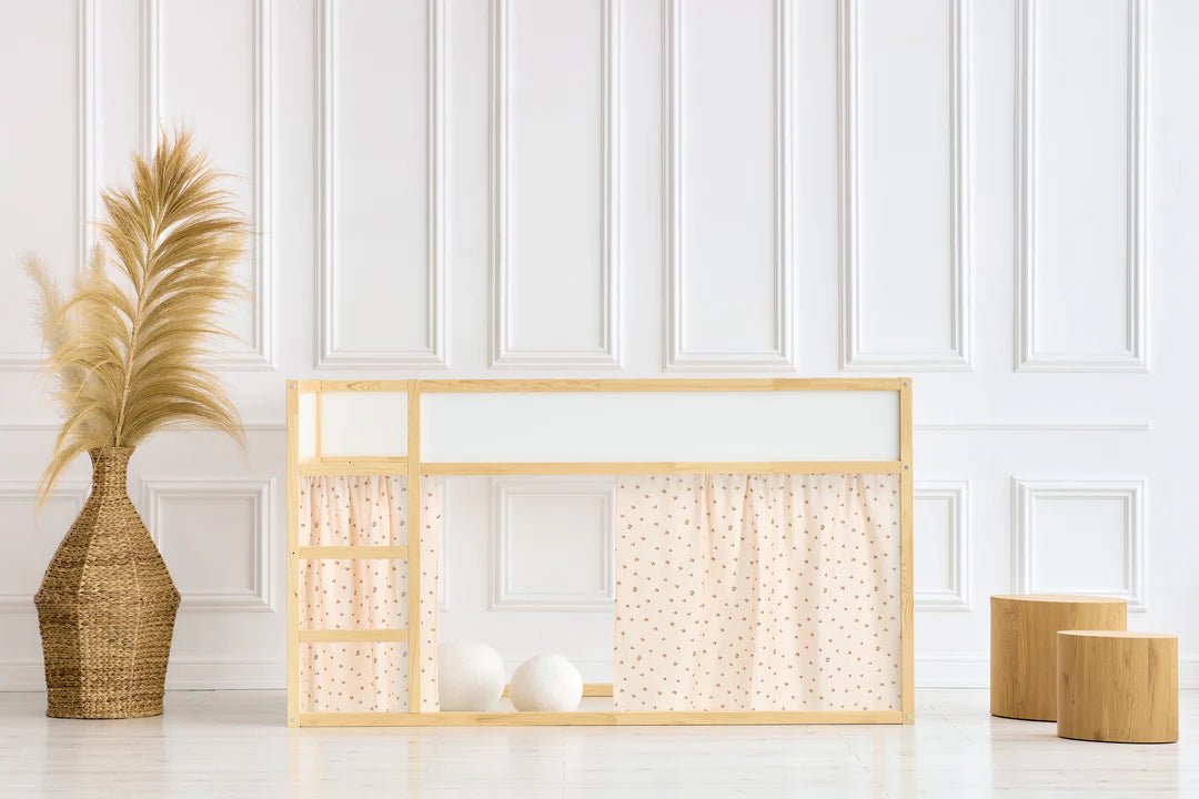 Minimalistisches Kinderspielhaus mit hellem Holzrahmen, gepunkteten Vorhängen und Stufen, neben einer großen Vase mit Pampasgras und zwei Holzhockern, vor einer weiß getäfelten Wand.