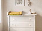 Ein KraftKids Wickelaufsatz in Weiß, passend für die 108 cm breite HEMNES Kommode (Wickelfläche 50 x 70 cm), mit gelber KraftKids Wickelauflage, gefalteten Handtüchern, Babyartikeln und Tierhaken unter Blattbildern in sanftem Licht.