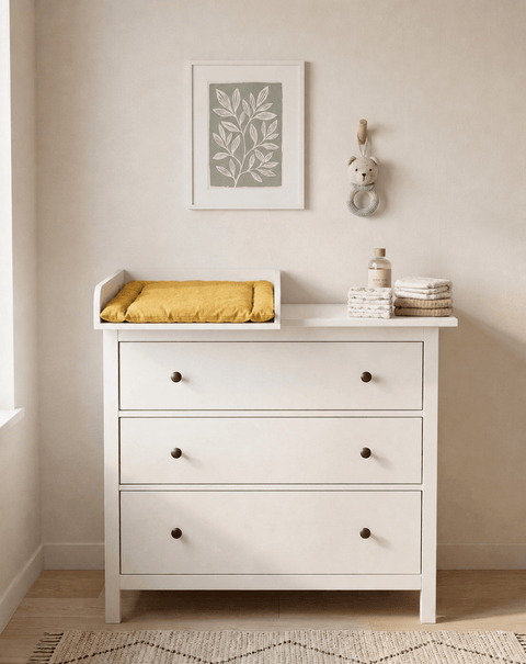 Ein KraftKids Wickelaufsatz in Weiß, passend für die 108 cm breite HEMNES Kommode (Wickelfläche 50 x 70 cm), mit gelber KraftKids Wickelauflage, gefalteten Handtüchern, Babyartikeln und Tierhaken unter Blattbildern in sanftem Licht.