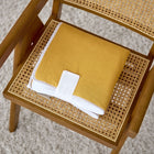 Eine gefaltete gelb-weiße Decke liegt auf einem Holzstuhl neben der KraftKids Reisewickelunterlage Leinen gelb senfgelb - ideal, damit Ihr Baby es zu Hause oder auf Reisen bequem hat.