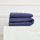 Ein ordentlich gefaltetes KraftKids Bettwäscheset Leinen blau dunkelblau mit weißer Paspel aus 100% Leinen steht auf einem weißen Sockel vor einer Wand mit Zierleiste.