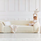 Ein kleines Mädchen sitzt auf einem cremefarbenen Sofa in einem hellen, minimalistischen Wohnzimmer und lächelt, während es das KraftKids Sternkissen Musselin Zweige braun über den Kopf hält. Pampasgras, Kissen und ein Korb akzentuieren diesen gemütlichen Raum - ideal für jedes Kinderzimmer.