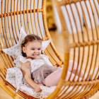 Ein kleines Mädchen mit geflochtenen Haaren sitzt lächelnd in einem großen Korbsessel mit weißen Kissen, trägt ein weißes Top und einen Tüllrock und genießt einen gemütlichen Moment in ihrem Kinderzimmer mit dem KraftKids Sternkissen Musselin Zweige braun.