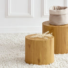 Zwei runde Holzhocker stehen auf einem weißen Zottelteppich. Auf dem einen steht das KraftKids Körbchen rund Musselin beige, ideal als Wickeltisch-Organizer, auf dem anderen liegen getrocknete Zierblätter. Im Hintergrund ist eine weiß getäfelte Wand zu sehen.