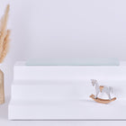 Ein weißer, dreistöckiger Ständer hält eine neuwertige Pusteblumen Sitzauflage für Ikea TROFAST von KraftKids GmbH, mit einer beigen Vase mit Pampasgras links und einem kleinen hölzernen Schaukelpferd unten rechts; der Hintergrund ist schlicht und hell.