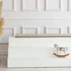 Ein minimalistisches Interieur mit weiß getäfelten Wänden, einer weißen Stufenbank mit der KraftKids GmbH Sitzauflage für Ikea TROFAST Musselin beige, einem dekorativen Holzschaukelpferd und einer hohen geflochtenen Vase mit getrocknetem Pampasgras.