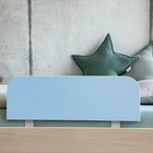 Ein modernes Kinderbett von KraftKids mit dem "Bettschutzgitter Rausfallschutz - blau hellblau", grünen sternförmigen Kissen, einem hellgrauen Kissen und Bettwäsche in sanften Pastellgrün- und Beigetönen vor einer Betonwand.