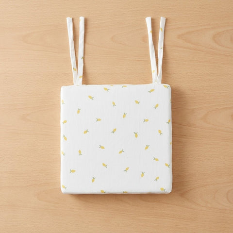 Das KraftKids Sitzkissen für Stühle aus Musselin ist ein weißes, quadratisches Stuhlkissen mit abnehmbarem Bezug, kleinen gelben Fruchtmotiven und zwei Paar Stoffbändern, das auf einer hellen Holzoberfläche abgebildet ist.