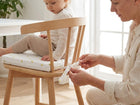 Ein Erwachsener bindet ein KraftKids Sitzkissen für Stühle aus Musselin an einen Holzstuhl, auf dem ein kleines Kind in einem hellen, stilvollen Esszimmer mit hellem Dekor sitzt, das Komfort und Design betont.