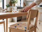 Eine Person stellt eine Schüssel auf einen hölzernen Esstisch mit Bechern, Holzutensilien und einer Serviette. Neben dem Tisch steht ein Stuhl mit dem KraftKids Sitzkissen für Stühle aus Musselin in einer hellen, modernen Küche.