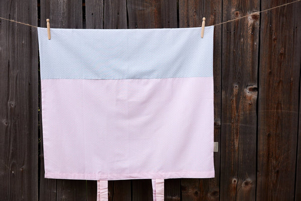 Der KraftKids Bezug für Wickeltischauflage kleine Blätter rosa auf Weiß aus 100% Baumwolle hängt an einer Wäscheleine vor einem Holzzaun. Der waschbare Bezug zeigt zwei rosa Bänder an der Unterkante.