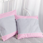 Zwei quadratische KraftKids Kissenbezüge Unigrau Unirosa mit hellgrauer Mitte und rosafarbener Umrandung ruhen vor einem weißen Metallkopfteil auf einer Zopfmusterdecke und ergänzen perfekt die elegante Bettwäsche und die weißen Vorhänge im Hintergrund.