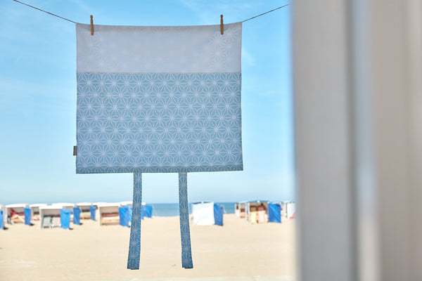 Der KraftKids Bezug für Wickeltischauflage weiße Diamante auf Pastell Blau (75 x 70 cm) hängt an einer Wäscheleine mit Bändern, vor einem sonnigen Strand mit Hütten und blauem Himmel - ideal als stilvoller Wickelauflagenbezug.