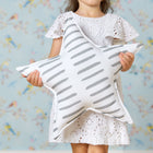 Ein kleines Kind in einem weißen Ösenkleid hält ein großes KraftKids Sternkissen graue Striche auf Weiß und steht vor einer pastellfarbenen, vogelgemusterten Tapete in einem gemütlichen Kinderzimmer.