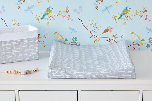 A KraftKids Bezug für Keilwickelauflage in Blau mit weißen Pfeilen liegt auf einer weißen Kommode neben passendem Korb und Schnullerkette, vor blauer Tapete mit bunten Vögeln und Libellen.