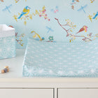 Ein Bezug für Keilwickelauflage weiße Diamante auf Pastell Blau von KraftKids aus 100% Baumwolle liegt auf einer weißen Kommode neben einem passenden Korb, umgeben von bunten Vögeln, Schmetterlingen und Zweigen sowie einer Schnullerkette.