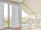 Helles, modernes Kinderzimmer mit großen Glastüren, KraftKids Gardinen Uniweiss Karo grau, hellem Holzboden, weißer Wiege und einem von der Decke hängenden Wolkenmobile. Sonnenlicht fällt ein und unterstreicht die klare, minimalistische Einrichtung.