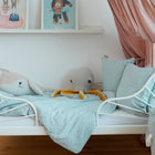 Ein weißes Metall-Kleinkinderbett mit dem KraftKids Bettwäscheset Musselin mint Pusteblumen, Plüschtieren mit Meerestieren, Kissen und einem rosa Baldachin. Gerahmte Kinderkunst befindet sich auf einem weißen Regal darüber, durch das Licht durch einen durchsichtigen Vorhang fällt.