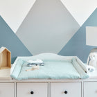 Eine weiße Kommode mit einer mintfarbenen Wickelauflage Musselin Pusteblumen von KraftKids, Babykleidung, eine Bürste, Spielzeughäuser mit Plüschtieren und eine Lampe mit einem Plüschhasen darauf stehen vor einer blau-weißen, geometrischen Wand.