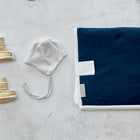 Ein Paar kleine beigefarbene Babyschühchen, eine grau gestreifte Babymütze mit Bändern, eine gefaltete marineblau-weiße Decke und die KraftKids Reisewickelunterlage Musselin dunkelblau liegen ordentlich auf einer hellgrauen Fläche.