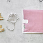 Ein Paar weiche hellbraune Babyschühchen, eine grau-weiß gestreifte Babymütze mit Bändern und eine rosa Decke mit weißem Rand auf einer hellen Betonfläche - perfekt mit Ihrer KraftKids Reisewickelunterlage Musselin rosa für unterwegs.