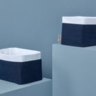 Zwei KraftKids Körbchen Musselin dunkelblaue Anker mit navyfarbenen Körpern und weißen Rändern sitzen auf hellblauen geometrischen Plattformen vor einem passenden Hintergrund - eine stilvolle Lösung für praktische Aufbewahrung am Wickeltisch.