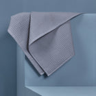 Eine KraftKids Babydecke Waffel Piqué grau ist über die Ecke eines hellblauen geometrischen Sockels drapiert, der vor einem passenden blauen Hintergrund steht.