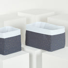 Zwei rechteckige KraftKids Körbchen Musselin goldene Punkte auf Grau, ideal für die Aufbewahrung auf dem Wickeltisch, werden auf weißen geometrischen Blöcken vor einem hellen Hintergrund dargestellt.