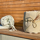 Zwei helle KraftKids Bettumrandung Nestchen Leinen Natur Braun Sets werden auf einer Holzfläche im Sonnenlicht ausgestellt - das eine gerollt und mit Schnur verschnürt, das andere lose gefaltet, mit einer Holzlattenwand als Hintergrund.