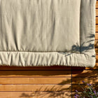 Eine KraftKids Krabbeldecke Leinen Natur Braun hängt über einer Holzwand im Sonnenlicht. Unten rechts sind Pflanzenschatten, lila Blumen und ein Leinen Körbchen für Wickeltisch sichtbar.