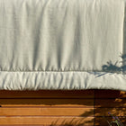 Eine Krabbeldecke Leinen leicht grünes Weiß von KraftKids hängt im Sonnenlicht über einem Holzzaun und wirft Schatten auf den Stoff und den Zaun. Pflanzenschatten sind sichtbar, mit einem Leinen Körbchen für Aufbewahrung in der Nähe in der unteren rechten Ecke.