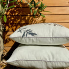 Zwei hellgrün-weiße Kissenbezüge Leinen von KraftKids sind im Freien auf Kies gestapelt, berührt von blattreichen Schatten. Ein Holzzaun mit Grünzeug schafft einen ruhigen Hintergrund für diese ruhige Bettwäscheszene.