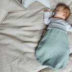 Ein Baby mit einem Schnuller schläft auf einem großen beigen Bett, eingewickelt in einen mintfarbenen KraftKids Strampelsack Herbst Winter Musselin, mit sternförmigen Kissen in der Nähe.