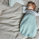 Ein Baby schläft auf einer beigen Bettgarnitur, gemütlich in einem KraftKids Strampelsack Herbst Winter Musselin mint Pusteblumen mit einem weißen Langarmshirt und Schnuller, umgeben von dekorativen sternförmigen Kissen.