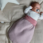 Ein Baby liegt mit einem Schnuller auf einem beigen Bettchen, eingehüllt in den rosa KraftKids Strampelsack Frühling Sommer Waffel Piqué. Große sternförmige Kissen liegen neben dem Baby und schaffen eine charmante und gemütliche Atmosphäre.