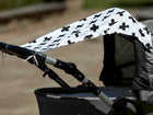 Ein grauer Kinderwagen wird an einem sonnigen Tag im Freien mit unscharfem Hintergrund von dem Sonnensegel schwarze Pluszeichen von KraftKids aus 100 % Baumwolle beschattet, das mit kräftigen schwarzen Pluszeichen versehen ist.