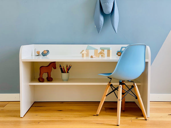 Der KraftKids Kinderschreibtisch weiß mit Zwischenboden steht auf einem Holzboden mit Holzspielzeug, Buntstiften in einer Tasse und einem blauen Stuhl vor einer blauen Wand, die mit einem ausgestopften Hasenkopf verziert ist.