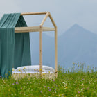 Ein schlichtes Holzbettgestell mit dem Bettbaldachin Musselin nilblau von KraftKids und weißem Bettzeug steht in einer Wildblumenwiese, im Hintergrund erheben sich neblige Berge unter einem wolkenverhangenen Himmel.