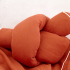 Die KraftKids Wickeltischumrandung Doppelkrepp Rot Herbstrot ist ein weiches, rostfarbenes Kissen mit weißer Paspelierung, das als verspielte Bettrolle gestaltet ist und auf einem passenden Stoff liegt.