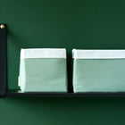 Zwei KraftKids Körbchen Doppelkrepp Grün Jade mit weißer Oberseite und jadegrünen Seiten sitzen auf einem schwarzen Wandregal - ideal zur Aufbewahrung auf oder über dem Wickeltisch vor einer dunkelgrünen Wand.