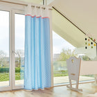 Ein helles Kinderzimmer mit großen Fenstern, hellen Holzböden und KraftKids Gardinen Uniweiss weiße Halbkreise auf Pastelblau aus 100% Baumwolle. Eine weiße Wiege steht neben einem modernen Mobile, und draußen sind Bäume und Häuser zu sehen.