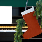 Eine KraftKids Weihnachtssocke Doppelkrepp Rot Herbstrot und eine Tannengirlande hängen an einer Metallleiter neben einem schwarzen Klavier mit Notenblättern. Der sanft verschwommene Hintergrund unterstreicht die festliche Weihnachtsdekoration.