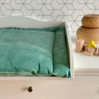 Eine KraftKids Wickelauflage Cord Breitcord Mint ruht auf einer weißen Kommode. Drei geflochtene Körbe sind in der Nähe gestapelt, und mehrere Holzklötze, einer davon mit einem gelben Dreieck, liegen vor einer geometrisch gemusterten Wand.