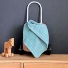 Ein Holzspielzeugpferd auf Rädern steht neben einem kleinen Weidenkorb mit schwarzen Rädern, einem weißen Griff und einer darüber drapierten mint Cord Breitcord Babydecke von KraftKids, alles auf hellem Holz vor einer dunklen Wand.