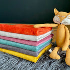 Ein Plüschfuchs sitzt auf einer grauen, pelzigen Unterlage neben bunten, gefalteten Decken und einer KraftKids Wickelunterlage Cord Breitcord in Reinorange, die alle vor einem dunklen Hintergrund angeordnet sind.