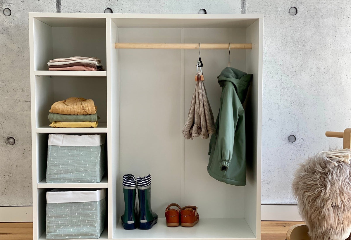 Der KraftKids Kleiderschrank für Kinder nach Montessori in Weiß bietet Regale mit gefalteter Kleidung, gemusterte Behälter, eine Stange mit einem Schal und einer grünen Jacke sowie Regenstiefel und Sandalen auf dem unteren Regal vor einer Betonwand.