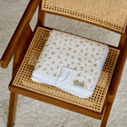 Eine gefaltete weiße Babydecke mit dezentem Blattgoldmuster ruht auf einem geflochtenen Rohrstuhl - ideal neben der KraftKids Reisewickelunterlage Musselin Zweige braun, alles auf einem hellen, strukturierten Teppich.